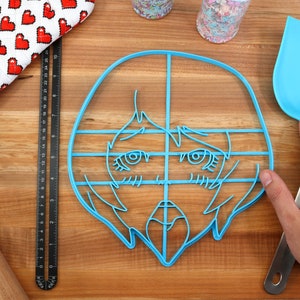 May include: A blue plastic cookie cutter shaped like an anime-style face with large eyes and a mouth. The cutter is on a wooden surface with a ruler, rolling pin, and spatula.