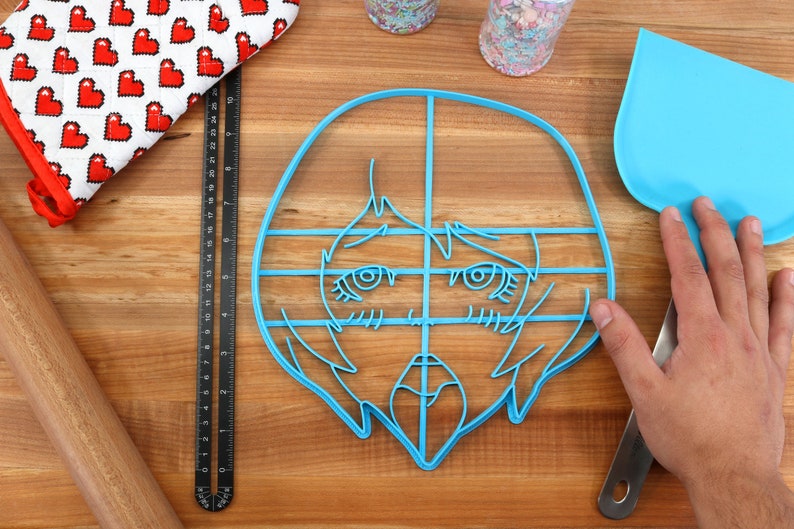 May include: A blue plastic cookie cutter shaped like an anime-style face with large eyes and a mouth. The cutter is on a wooden surface with a ruler, rolling pin, and spatula.