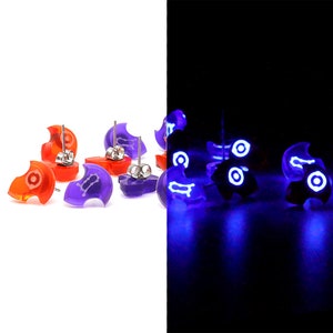 May include: A set of glow-in-the-dark earrings. The earrings are shaped like a ghost and are purple and orange. The earrings glow blue in the dark.