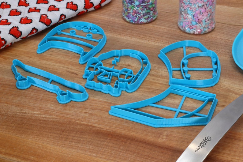 May include: A set of five blue plastic cookie cutters shaped like a skateboard, a skull, a hat, a surfboard, and a pair of headphones.