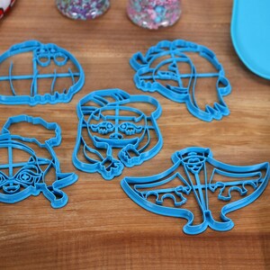 May include: Five blue plastic cookie cutters shaped like cartoon characters. The cutters are on a wooden surface.