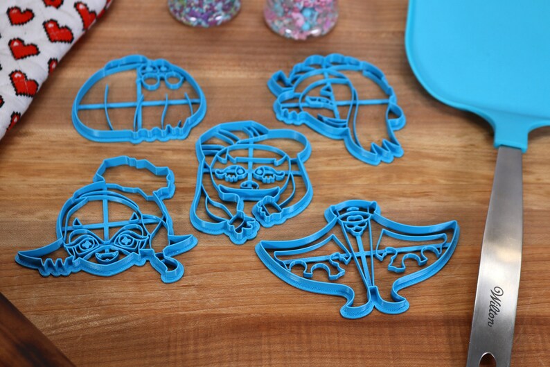 May include: Five blue plastic cookie cutters shaped like cartoon characters. The cutters are on a wooden surface.