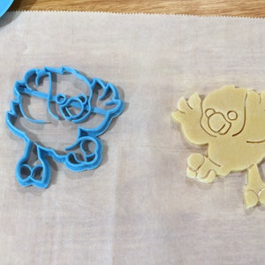 May include: A blue plastic cookie cutter and a beige cookie in the shape of a bird. Both are on a white parchment paper surface.