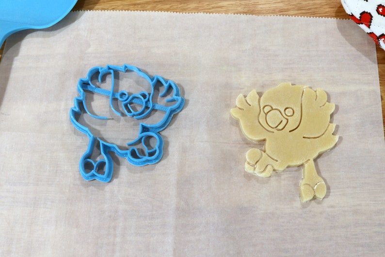 May include: A blue plastic cookie cutter and a beige cookie in the shape of a bird. Both are on a white parchment paper surface.