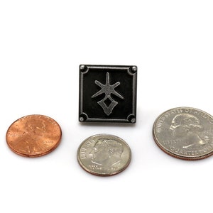 May include: A black metal pin with a stylized symbol in the center. The pin is about the size of a US quarter.