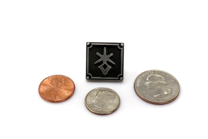 May include: A black metal pin with a stylized symbol in the center. The pin is about the size of a US quarter.