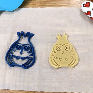 May include: Two cookie cutters, one blue and one beige, shaped like a cartoon owl with a crown on its head. The blue cutter is on a white parchment paper surface.