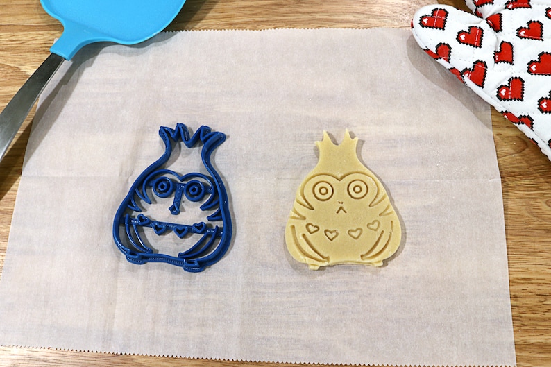 May include: Two cookie cutters, one blue and one beige, shaped like a cartoon owl with a crown on its head. The blue cutter is on a white parchment paper surface.