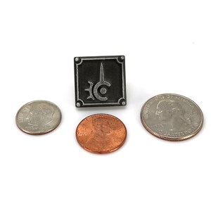 May include: A black and silver metal pin with a stylized sword and circle design. The pin is about the size of a US quarter.