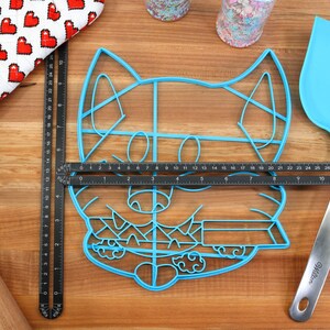 May include: A blue plastic cookie cutter shaped like a cartoon dog's face. The cutter is on a wooden surface with a ruler and a spatula.