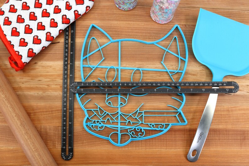 May include: A blue plastic cookie cutter shaped like a cartoon dog's face. The cutter is on a wooden surface with a ruler and a spatula.
