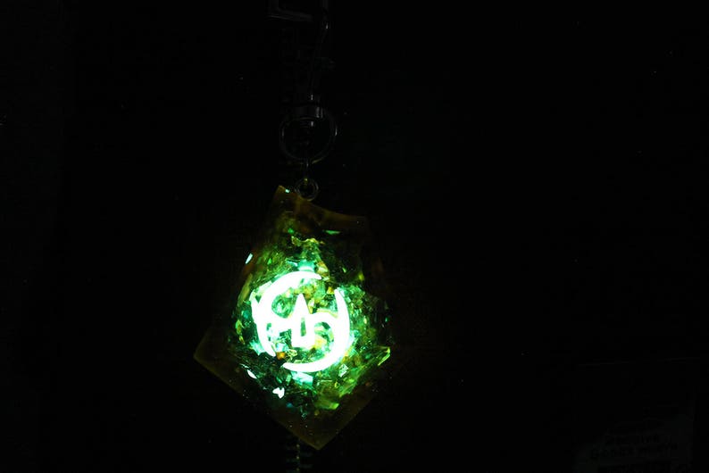 May include: A green and yellow resin pendant with a glowing white symbol inside. The pendant is on a black chain.