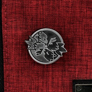 Op de afbeelding: Een zilveren metalen pin met een zwart-wit ontwerp van een gestileerde bloem met drie bloemblaadjes. De pin is op een rode stoffen achtergrond.