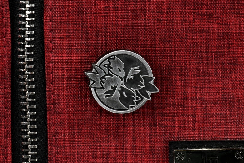 Op de afbeelding: Een zilveren metalen pin met een zwart-wit ontwerp van een gestileerde bloem met drie bloemblaadjes. De pin is op een rode stoffen achtergrond.