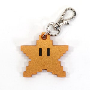 May include: A gold star-shaped keychain with a pixelated design and black eyes. The keychain has a silver metal loop.