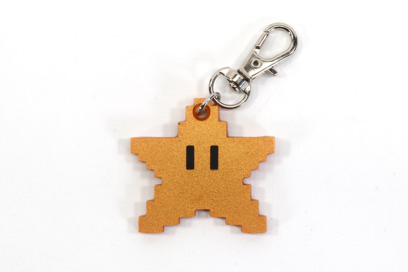 May include: A gold star-shaped keychain with a pixelated design and black eyes. The keychain has a silver metal loop.