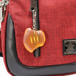 May include: A red crossbody bag with a black leather trim and silver zippers. A clear, amber-colored charm hangs from the zipper. The bag has a small metal plaque with text.