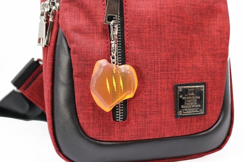 May include: A red crossbody bag with a black leather trim and silver zippers. A clear, amber-colored charm hangs from the zipper. The bag has a small metal plaque with text.