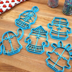 May include: A set of five blue plastic cookie cutters shaped like cartoon characters. The cutters are on a wooden surface.