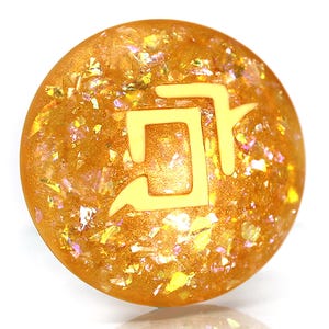 May include: A round, gold-colored resin orb with a light yellow symbol embedded inside. The orb is covered in iridescent glitter.