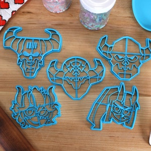May include: Five blue plastic cookie cutters shaped like cartoon demon heads. The cutters are on a wooden surface.