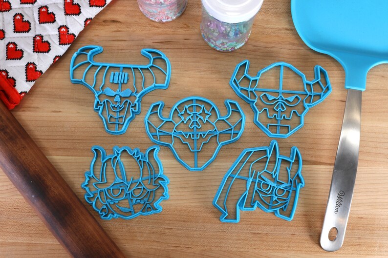May include: Five blue plastic cookie cutters shaped like cartoon demon heads. The cutters are on a wooden surface.