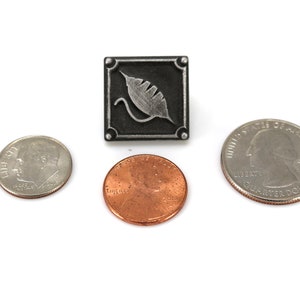 May include: A silver-colored metal pin with a black square background and a silver leaf design. The pin is shown next to a US quarter and a US penny for size comparison.
