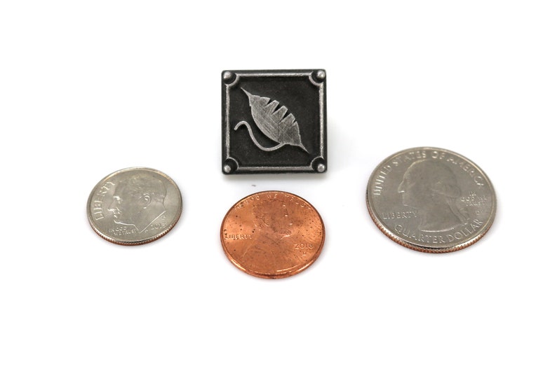 May include: A silver-colored metal pin with a black square background and a silver leaf design. The pin is shown next to a US quarter and a US penny for size comparison.