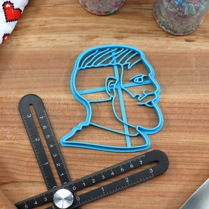 Op de afbeelding: Een blauwe plastic koekjesvormer in de vorm van een mensenhoofd met een baard en een lijn over de ogen. De koekjesvormer ligt op een houten oppervlak met een liniaal en een deegroller.