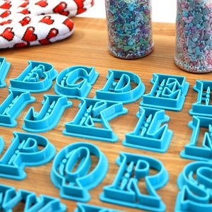 May include: A set of blue plastic alphabet cookie cutters on a wooden surface. The cutters are in a serif font style.