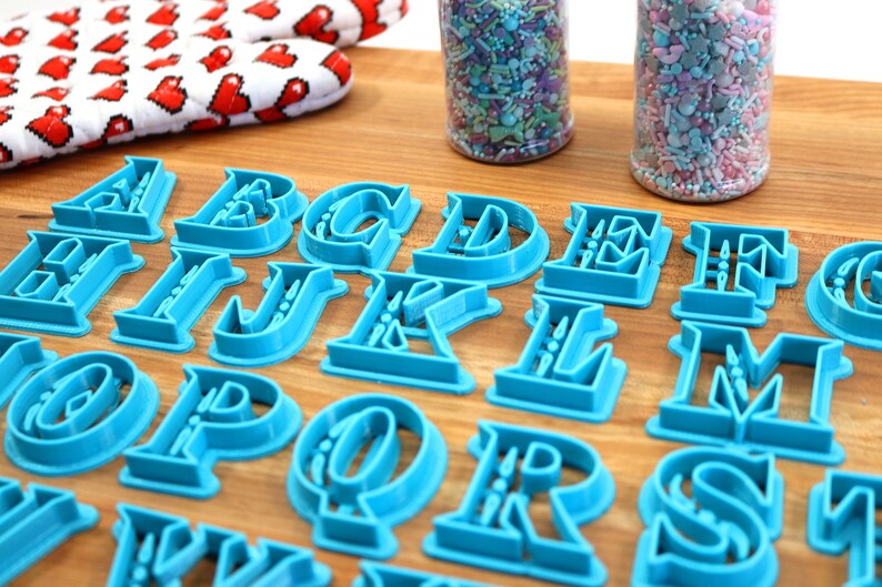 May include: A set of blue plastic alphabet cookie cutters on a wooden surface. The cutters are in a serif font style.