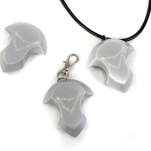 May include: Three silver-colored pendants with a white design in the shape of a constellation. One pendant is on a black cord necklace, one is on a silver keychain, and one is laying on a white surface.