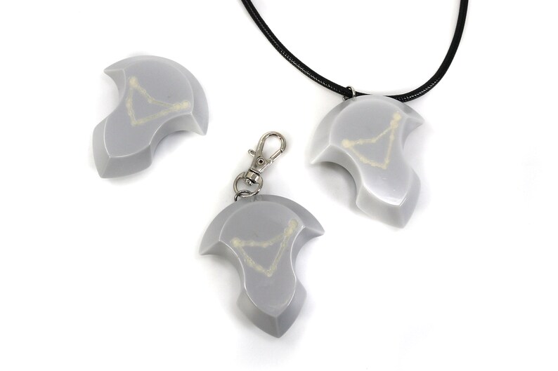 May include: Three silver-colored pendants with a white design in the shape of a constellation. One pendant is on a black cord necklace, one is on a silver keychain, and one is laying on a white surface.