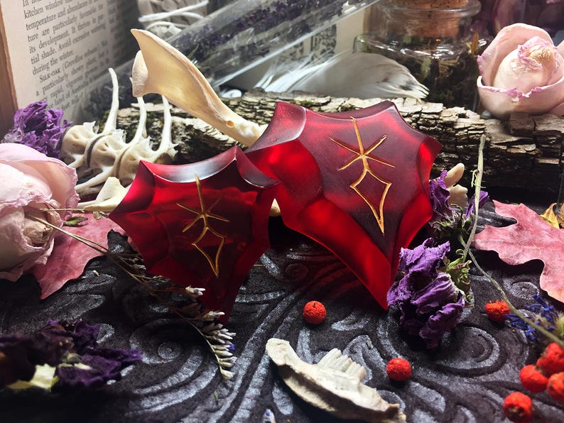 May include: Two red resin heart-shaped pendants with gold-colored symbols on a dark background. The pendants are surrounded by dried flowers and other natural elements.