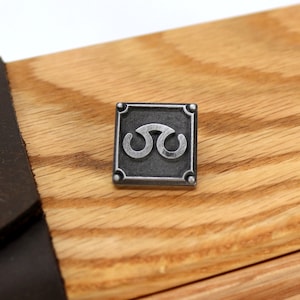 May include: A silver metal pin with a square frame and a stylized symbol inside. The symbol is a double-loop design with a silver finish.