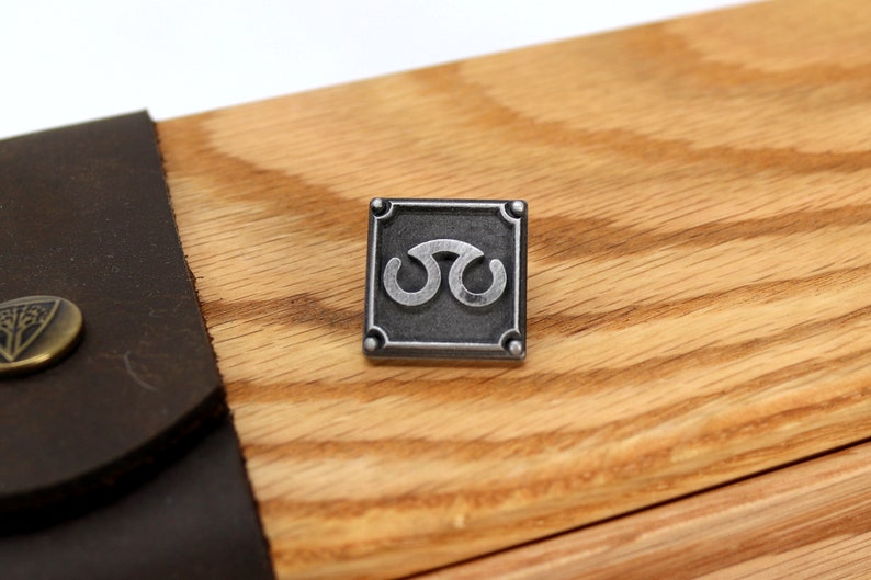 May include: A silver metal pin with a square frame and a stylized symbol inside. The symbol is a double-loop design with a silver finish.