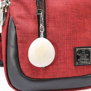 May include: A red crossbody bag with a black zipper and a white oval-shaped charm attached to the zipper. The bag has a black leather trim and a silver tag that reads "Where is the heart to receive good ware".