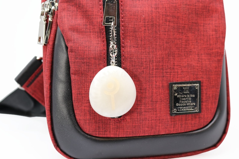 May include: A red crossbody bag with a black zipper and a white oval-shaped charm attached to the zipper. The bag has a black leather trim and a silver tag that reads "Where is the heart to receive good ware".