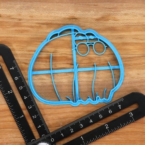 May include: A blue plastic cookie cutter shaped like a cartoon character wearing glasses. The cookie cutter has a detailed design with lines and curves.