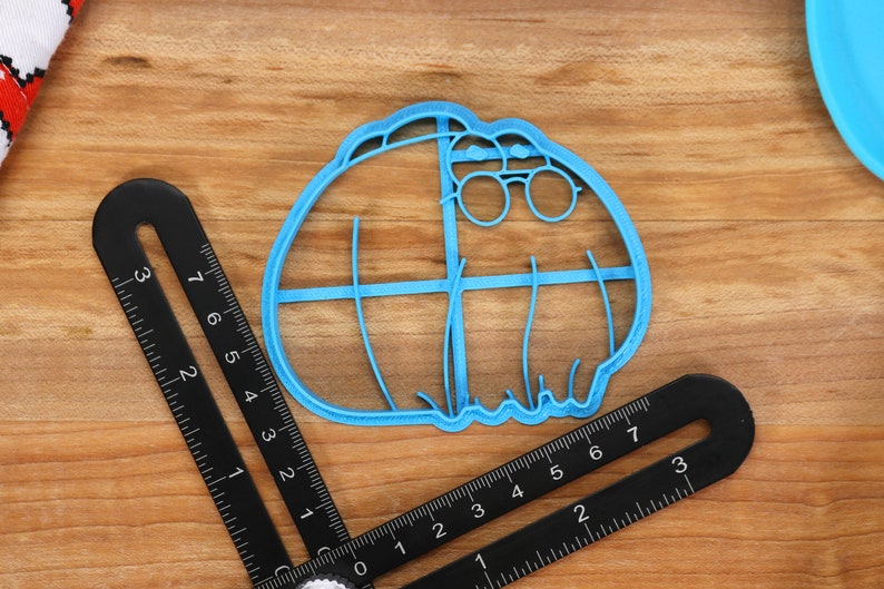 May include: A blue plastic cookie cutter shaped like a cartoon character wearing glasses. The cookie cutter has a detailed design with lines and curves.