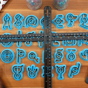 May include: A set of 26 blue plastic cookie cutters shaped like letters of the alphabet. The cutters are arranged in alphabetical order on a wooden surface.