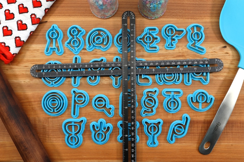 May include: A set of 26 blue plastic cookie cutters shaped like letters of the alphabet. The cutters are arranged in alphabetical order on a wooden surface.