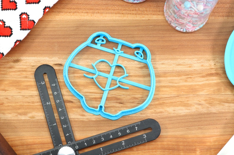 May include: A light blue plastic cookie cutter shaped like a cartoon bear with a heart in its chest. The cutter is on a wooden surface.