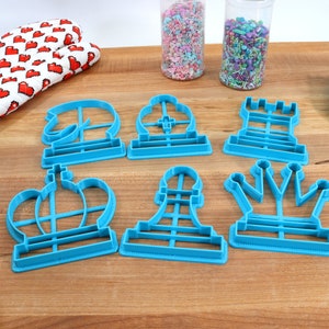 May include: A set of six blue plastic cookie cutters shaped like chess pieces: a king, queen, rook, bishop, knight, and pawn. The cutters are on a wooden surface with sprinkles in jars in the background.