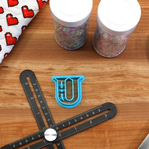 May include: A blue plastic cookie cutter shaped like the letter U, on a wooden surface with a black ruler, two jars of sprinkles, a blue spatula, and a red and white heart-patterned kitchen towel.