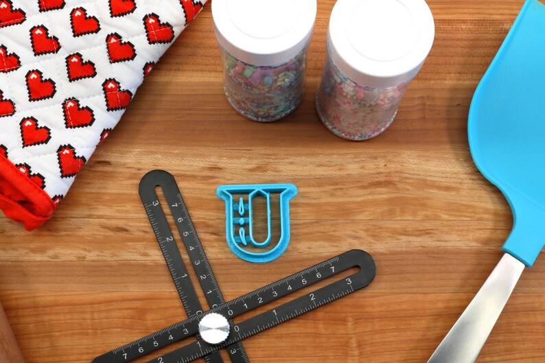 May include: A blue plastic cookie cutter shaped like the letter U, on a wooden surface with a black ruler, two jars of sprinkles, a blue spatula, and a red and white heart-patterned kitchen towel.