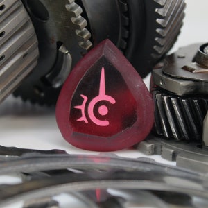 May include: A close-up of a red, heart-shaped, resin guitar pick with a pink symbol on it. The pick is sitting on top of a pile of metal gears.