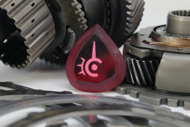 May include: A close-up of a red, heart-shaped, resin guitar pick with a pink symbol on it. The pick is sitting on top of a pile of metal gears.