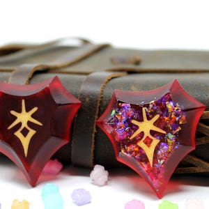 May include: Two red resin heart-shaped charms with gold glitter and a gold symbol in the center. The charms are sitting on a brown leather journal.