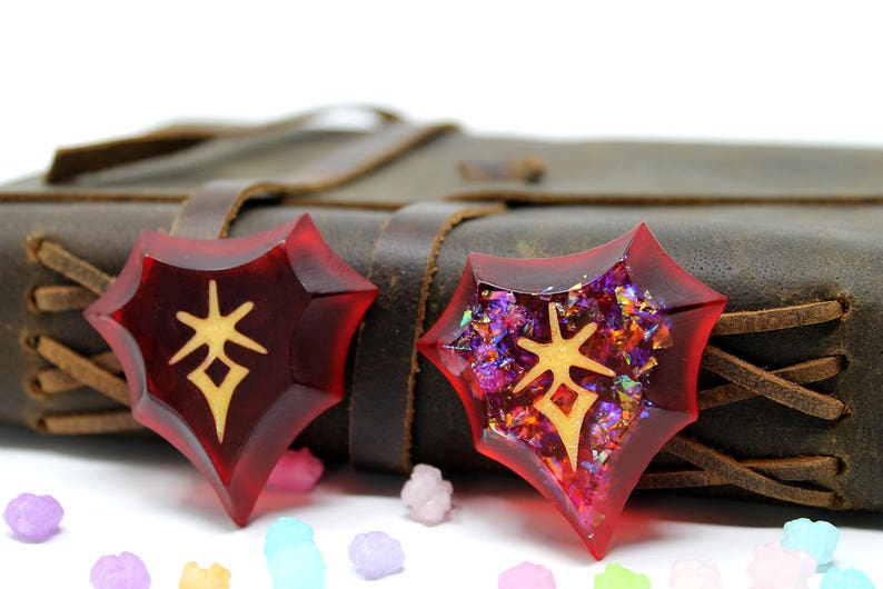 May include: Two red resin heart-shaped charms with gold glitter and a gold symbol in the center. The charms are sitting on a brown leather journal.